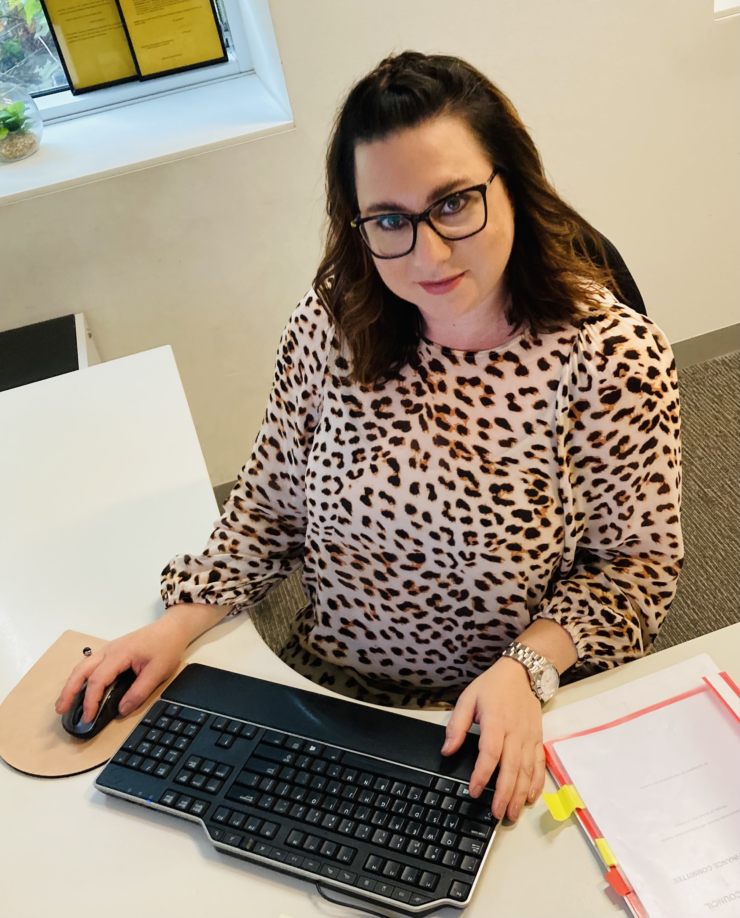 Angela is wearing her brunette hair in a half-up half-down style. She is wearing black framed glasses and a leopard print blouse. On her right hand is a silver watch and next to her hand is a computer mouse and keyboard. She is looking up at the camera smiling.