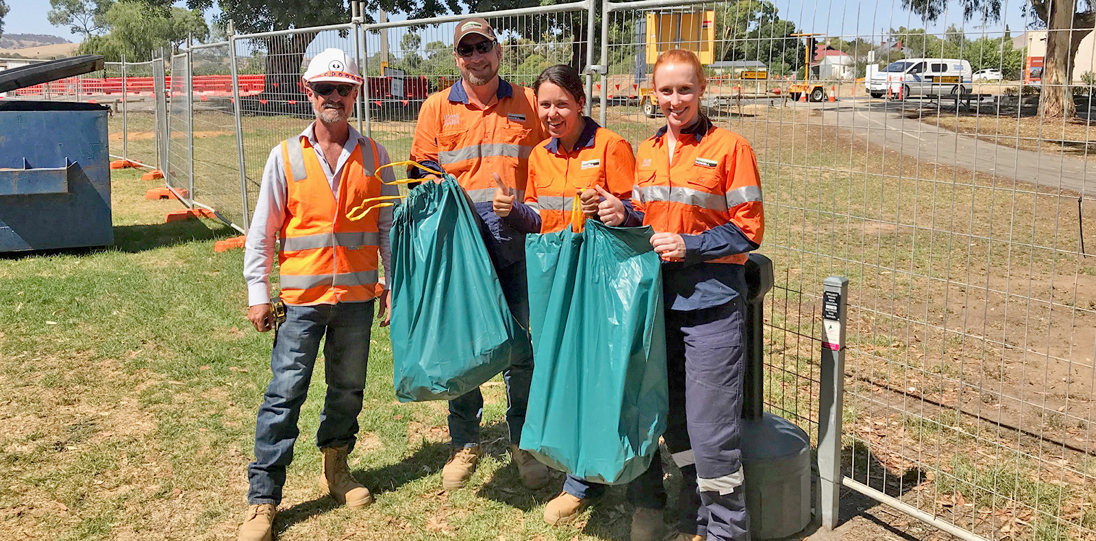 Four DPTI staff wearing hi-vis clothing are holding partially filled rubbish bags. There are two men on the left and two women on the right and they are standing in front of a rectangular mesh wire fence with a worksite behind it.