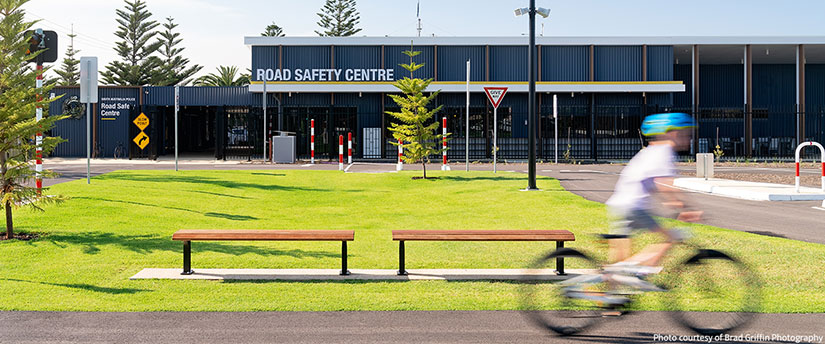 A cyclist rides past the Road Safety Centre building, surrounded by green grass and trees.