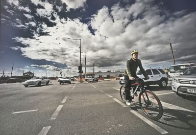 A man riding a bike across an intersection next to the construction site. There are cars on either side and an empty building surrounded by fencing in the background.