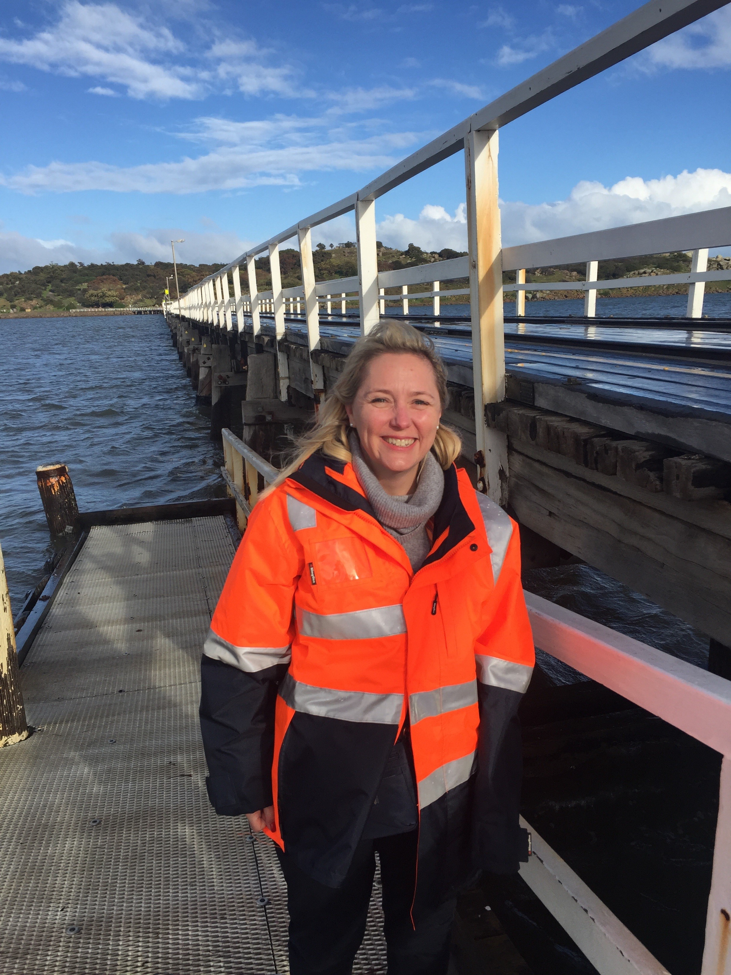 Emma Kokar is wearing an orange high vis jacket and is standing by the water on a jetty. Emma has mid length blonde hair and is smiling