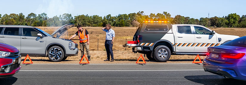 Two men stand by a broken-down car on the side of a road. A breakdown service vehicle with flashing amber lights is parked behind it. Warning triangles are on the road.