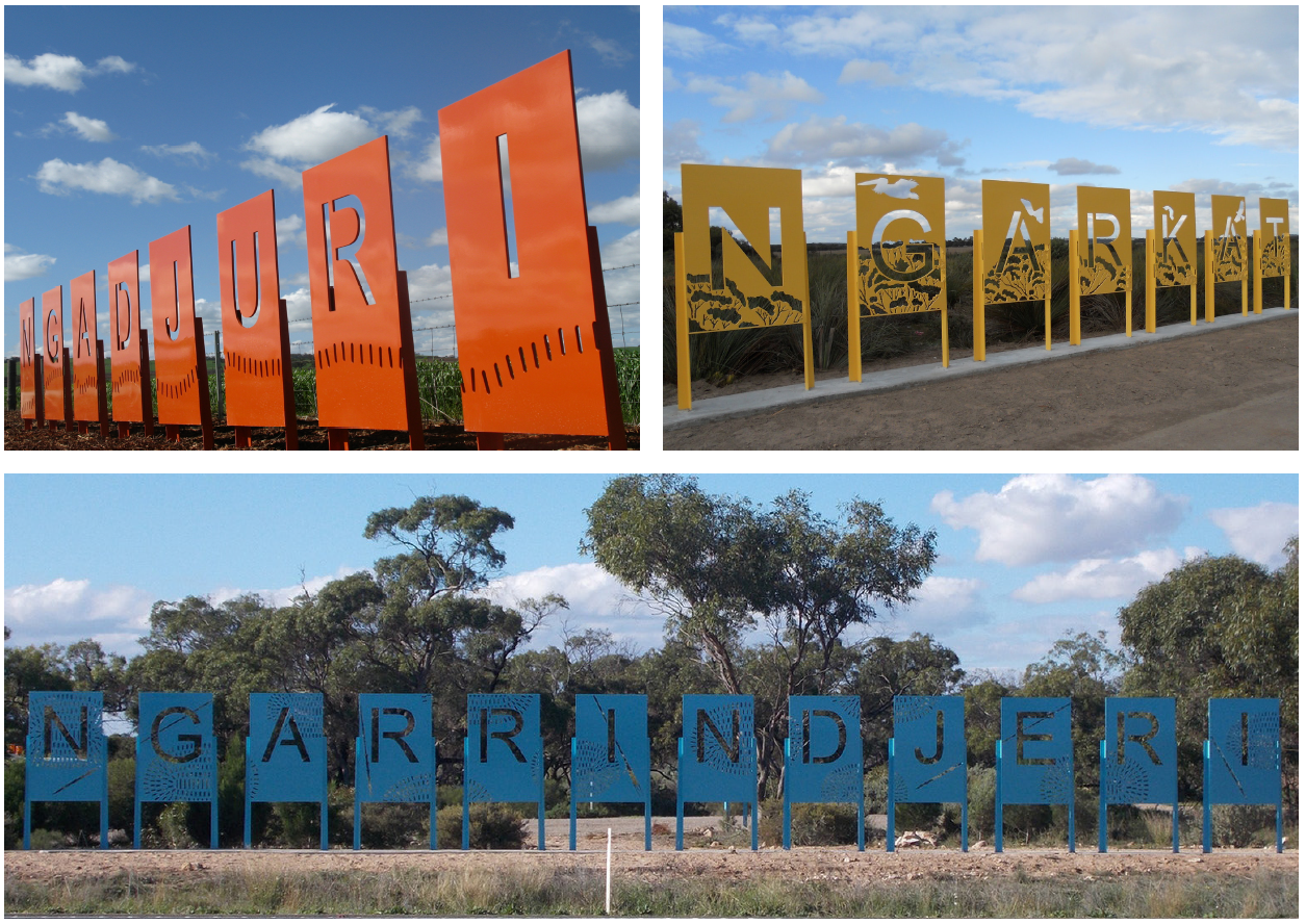 Three of the road signs, two placed next to each other abover a wider one below. The top left is orange in colour and spells out NGADJURI. On Each of the signs , each letter is etched out of a rectangular piece of metal and Aboriginal artwork surrounds it. The second sign is yellow in colour and spelss out NGARKAT. The rhird sign is blue in colour and says NGARRINDJERI.