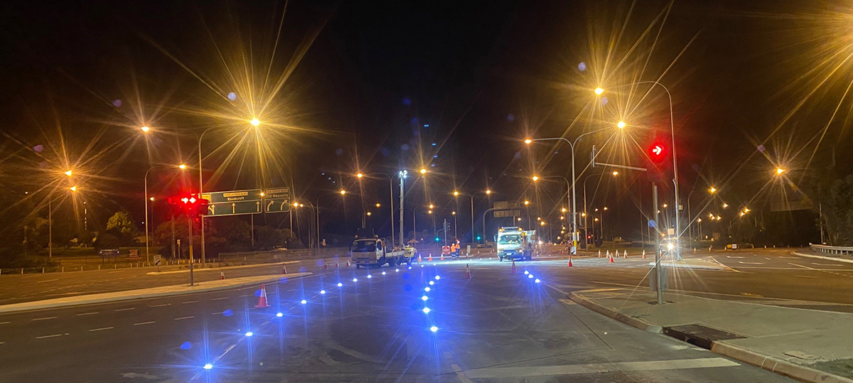An intersection at night with bright LED lights embedded in the road, showing the lane markings. There are road work crews and trucks with safety cones surrounding them in the background.