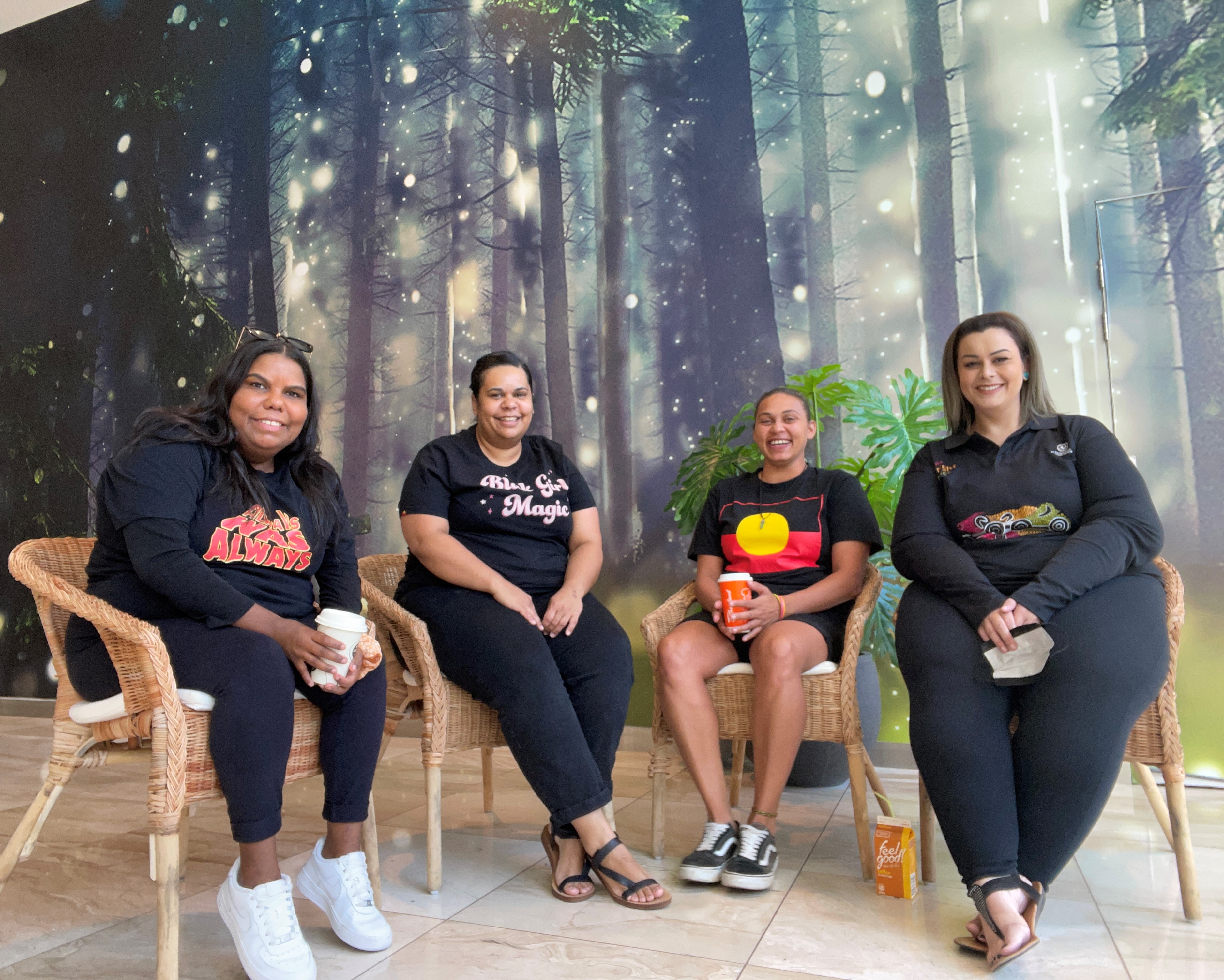 Four women, wearing black pants and a variety of Indigenous artwork tshirts shit in a row with big smiles. They are sitting in wooden chairs in front of a decorative painted wall