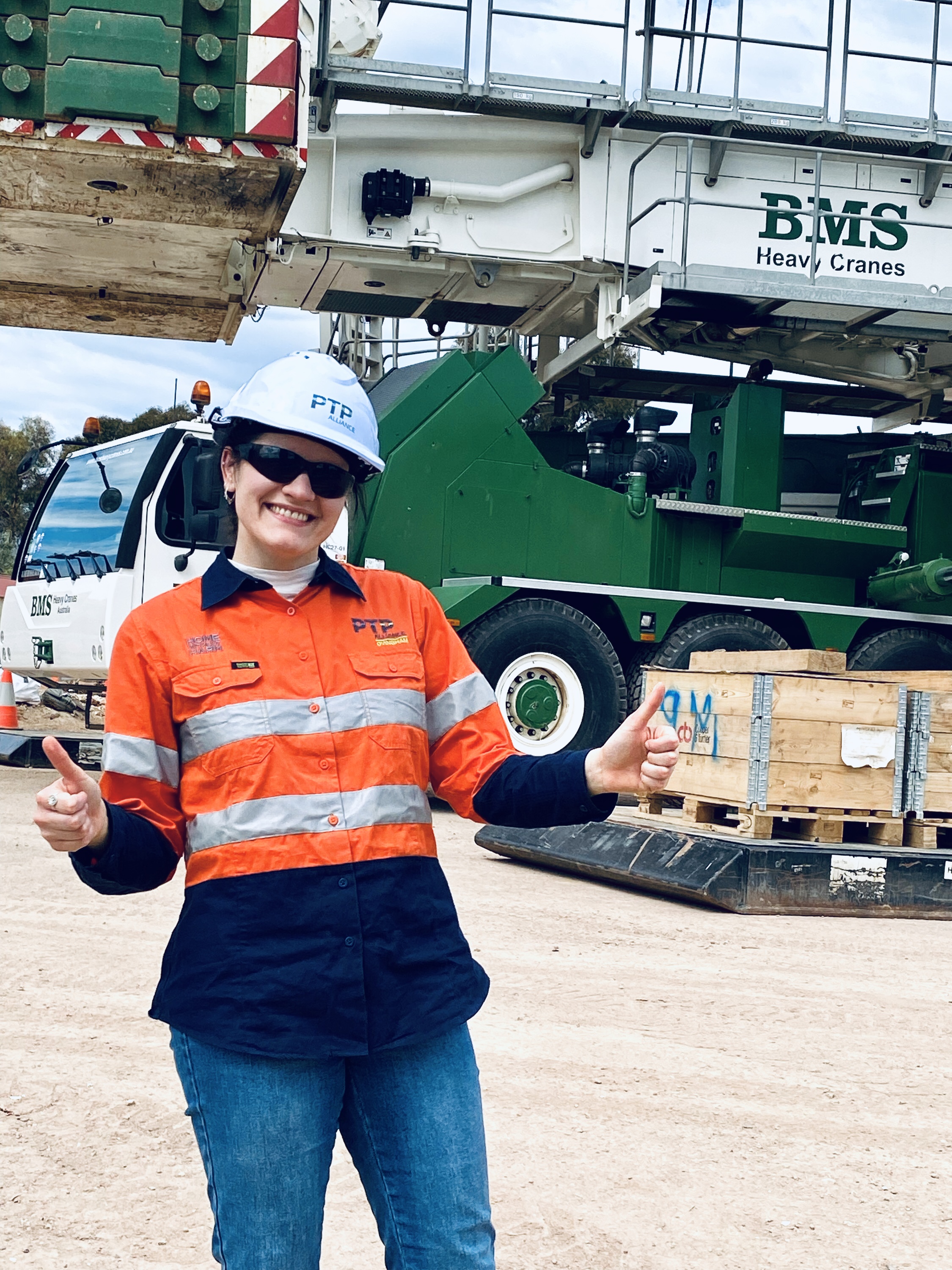 Teodara is wearing a white helmet and orange high vis long sleeved shirt. She is standing behind work site with a truck. She is smiling and is posing with two thumbs up