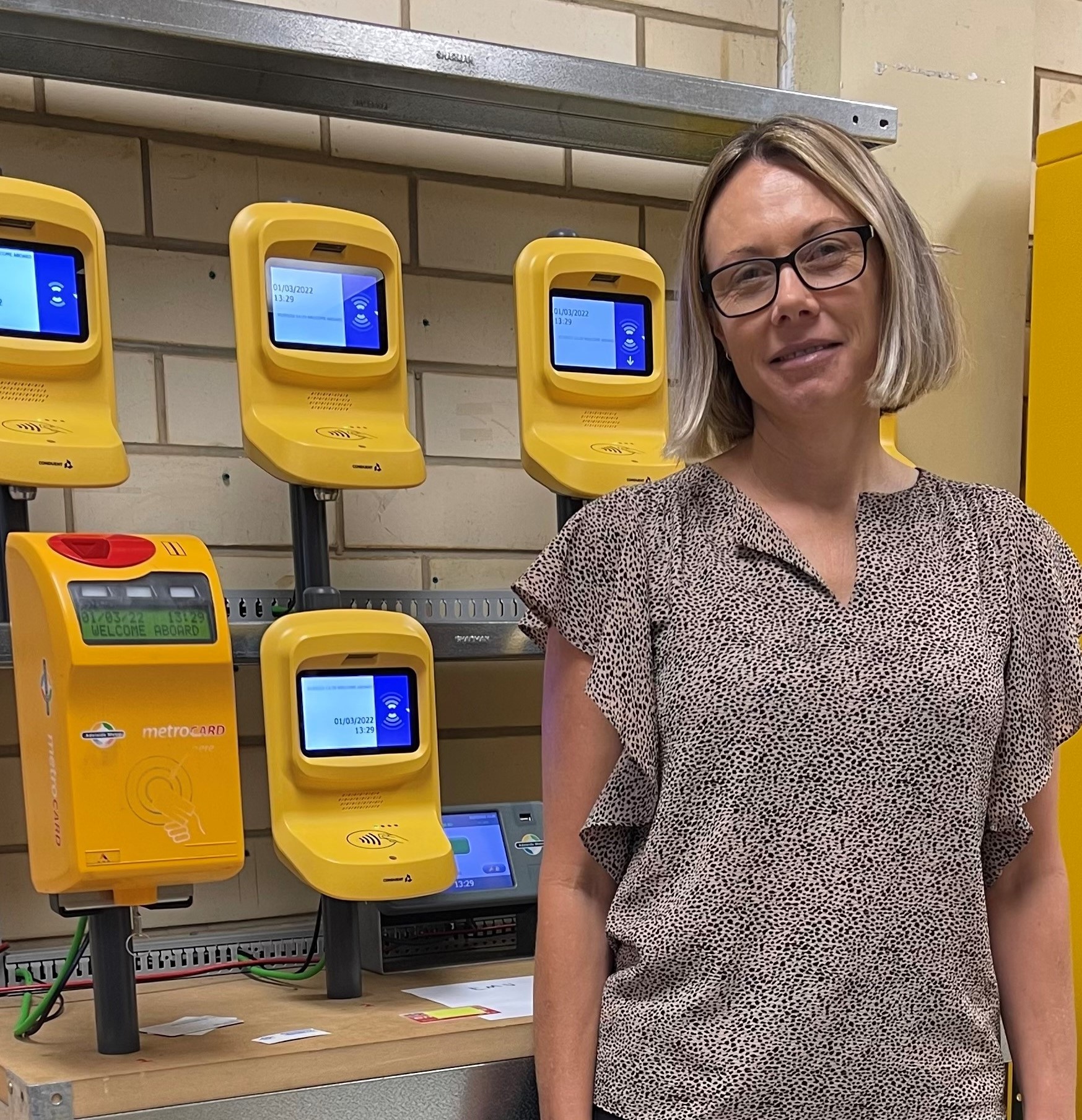 Tammy has blonde hair which is cut to about-chin length. She is wearing dark framed glasses and is wearing a brown office shirt. She is standing infront of yellow Adelaide Metro ticketing machines