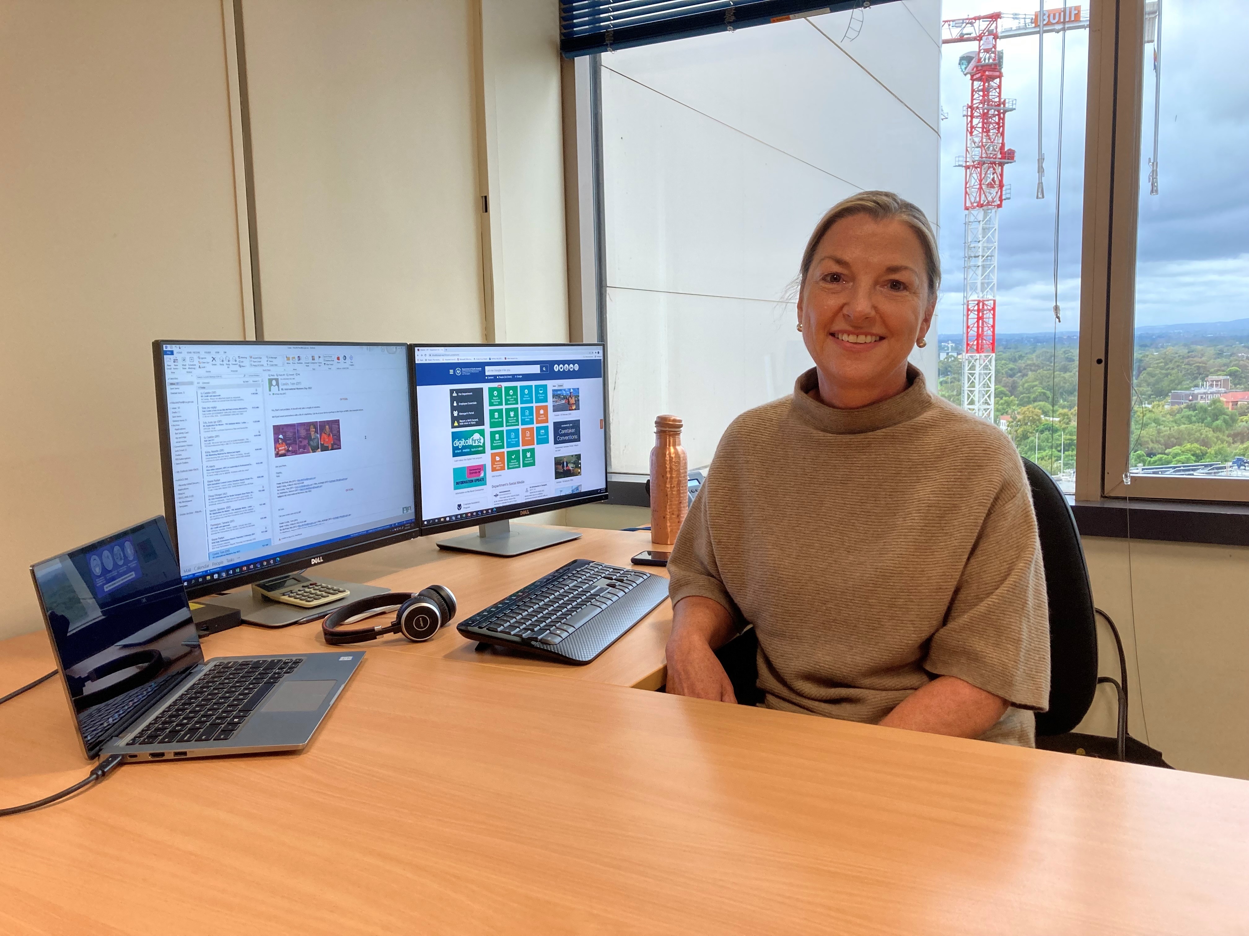 Rita is sitting in a bright office with two computer screens and a laptop behind her. Her office fronts a crane working on a project in the background. She is wearing a brown sweater and her blonde hair is tied back behind her ears. Rita is smiling