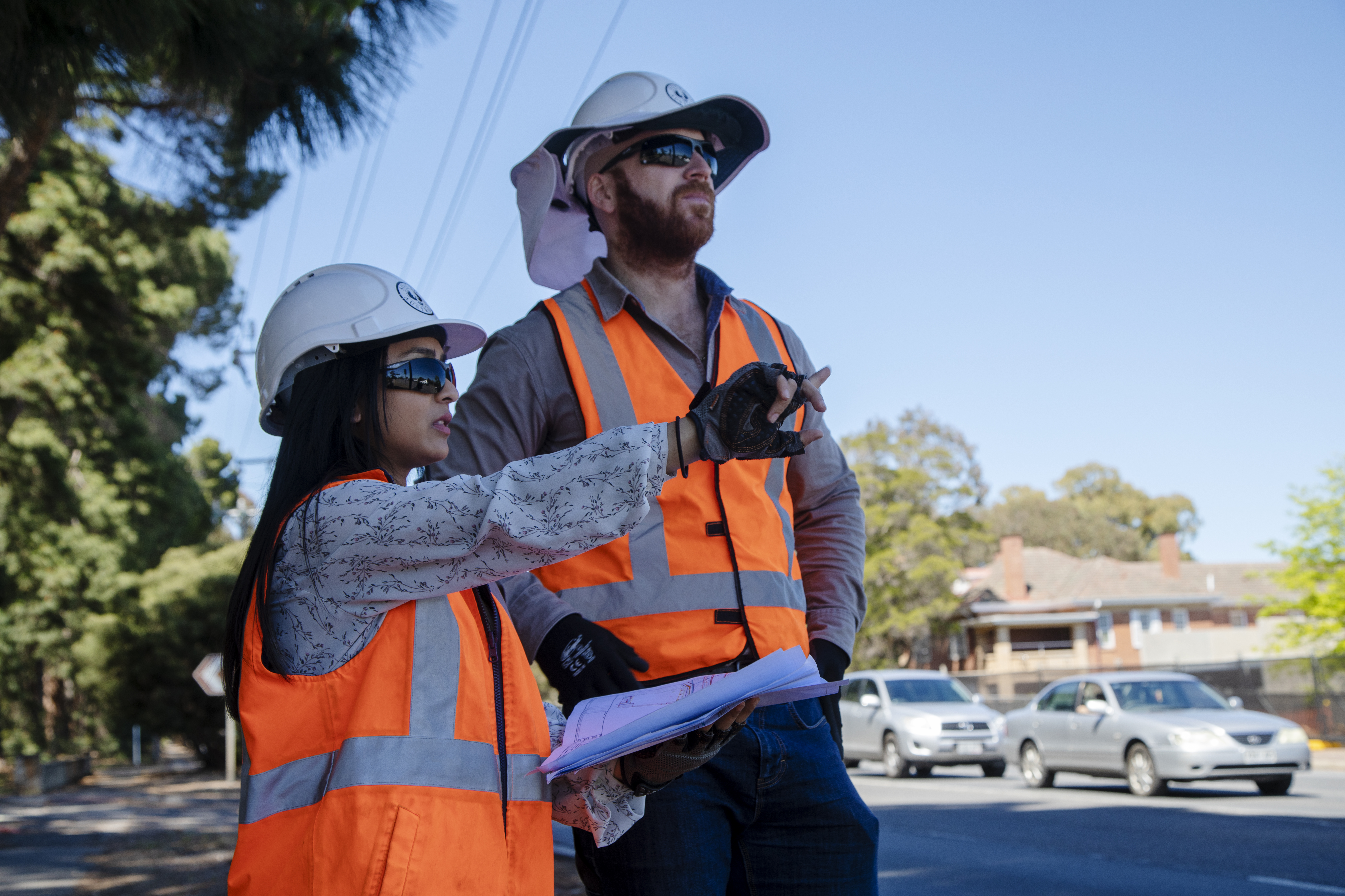 Vini is wearing an orange high vis vest, black sunglasses and a white safety helmet. She is standing next to a man with a beard who is also wearing an orange vest and white helmet. Vini is holding paperwork and pointing into the distance