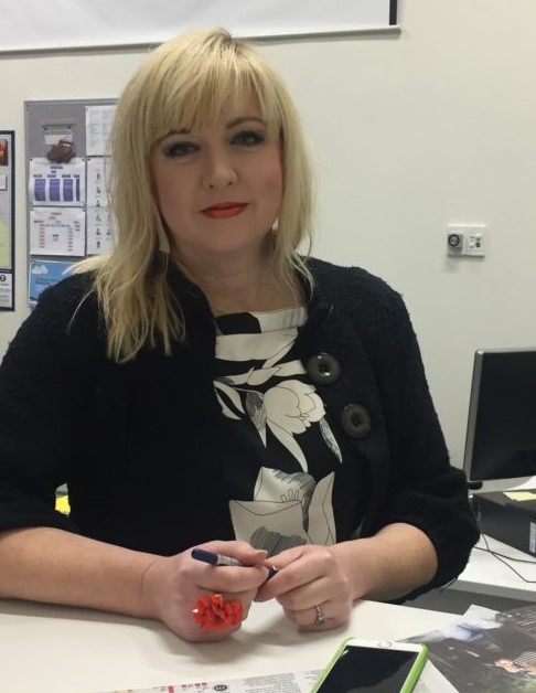 Paula is sitting behind a desk holding a black pen. She has short blonde hair with a front fringe and is wearing red lipstick. She is wearing a black blazer with a black and white floral print shirt underneath.