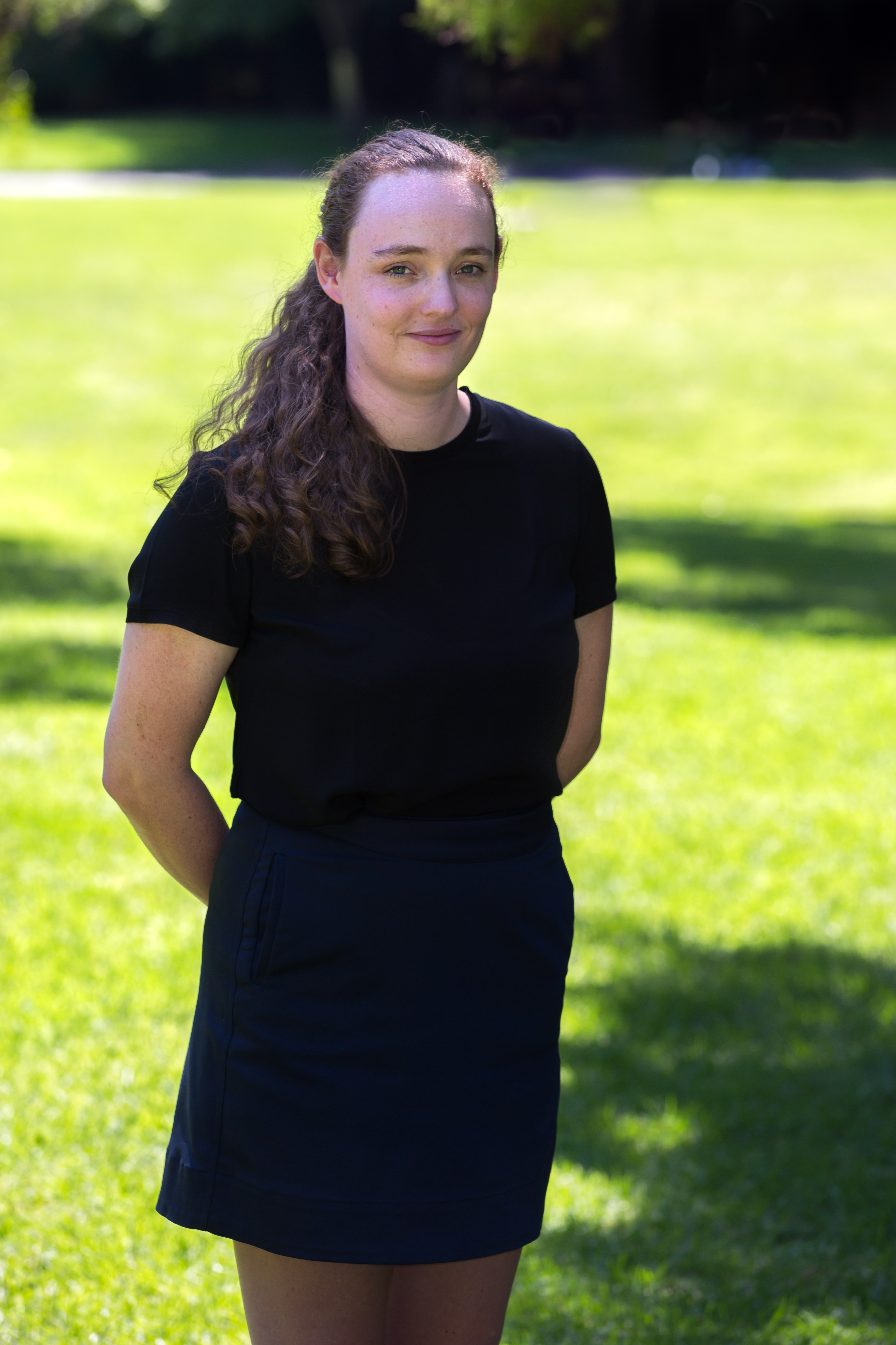 Arna is wearing a black tshirt and skirt, standing with her hands behind her back in a park. Her brown hair is tied into a low ponytail.